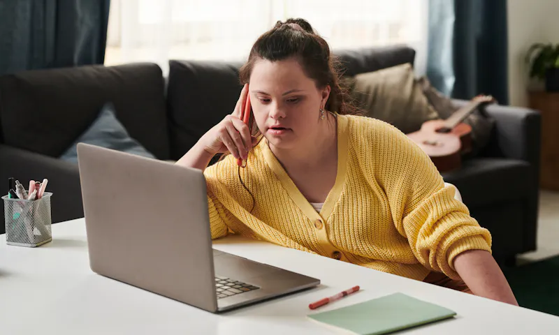 A young woman is seated in a lounge room. She's at a desk with her laptop open, chatting on the phone. She rests her elbow on the table and is wearing a yellow cardigan.