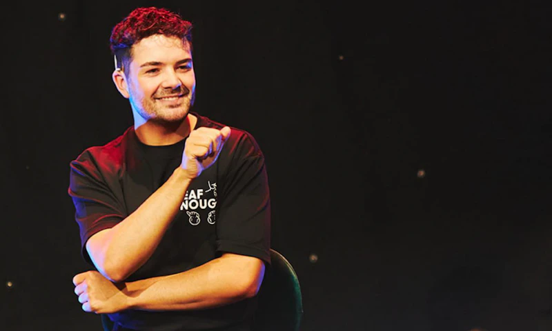 Nathan Borg, a deaf actor, sits beneath a spotlight on stage. He's signing in Auslan and wearing a black top that reads 'Deaf Enough'.