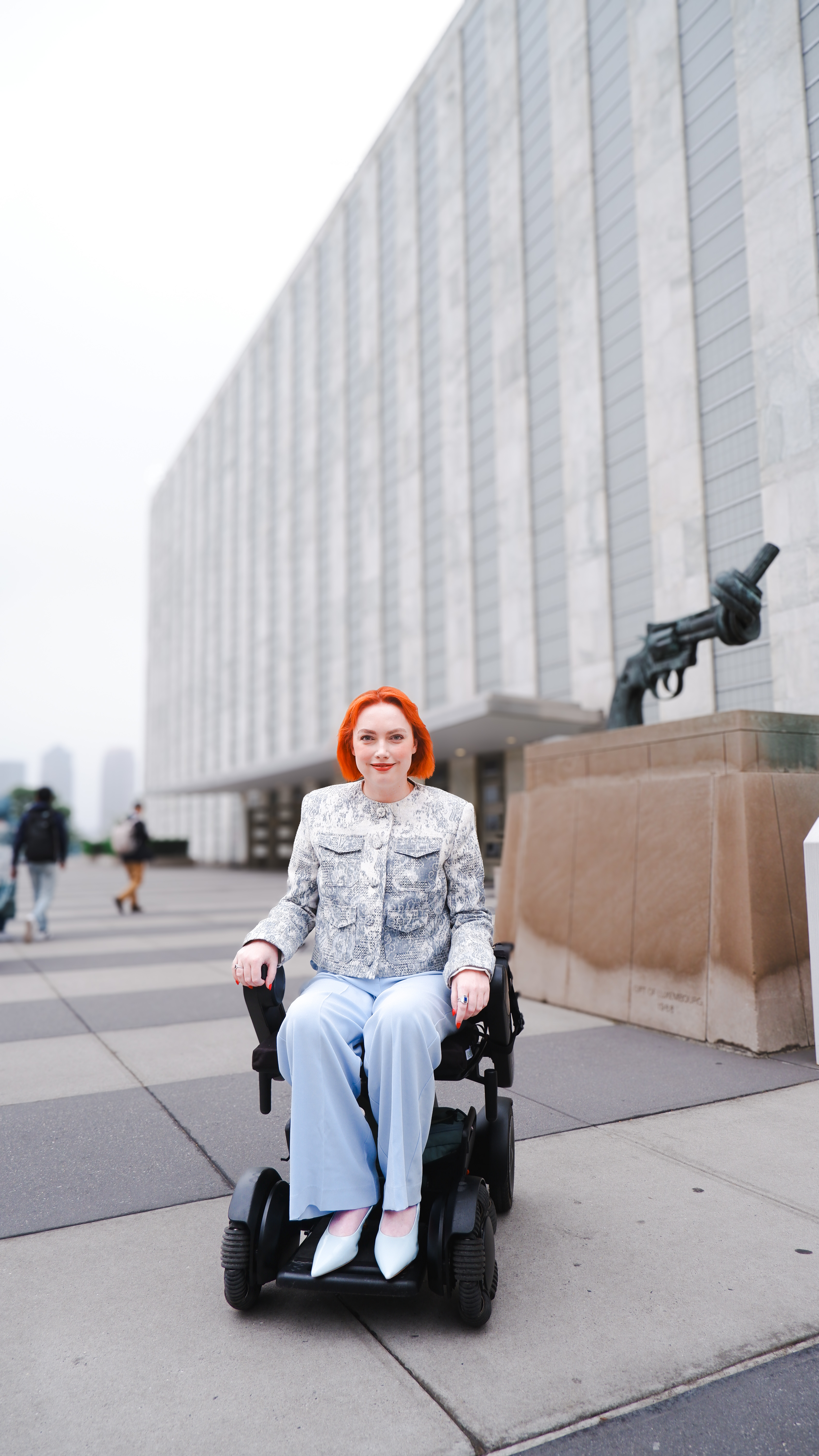 Elly Desmarchelier is outside the iconic United Nations building - a long rectangular building made up of windows and concrete. She is dressed in a grey suit jacket and blue trousers. Elly is a wheelchair user.