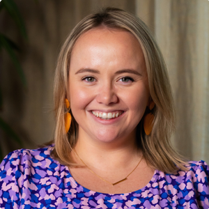Lizzy is smiling. She has short blonde hair and is dressed in a purple polka dot top, complemented by orange earrings.