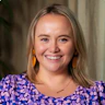 Lizzy is smiling. She has short blonde hair and is dressed in a purple polka dot top, complemented by orange earrings.