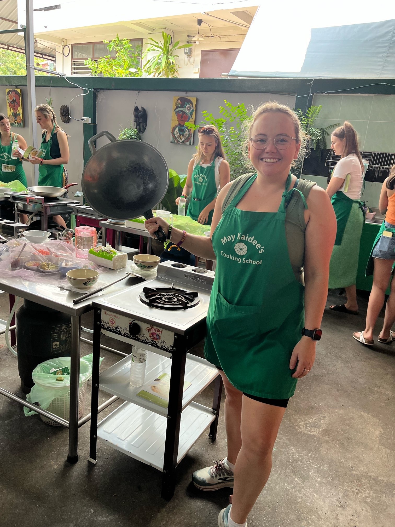 At an outdoor cooking class, Lizzy is holding up a wok. She's wearing a green apron and is smiling. Fellow cooking students are working at their cooking stations.