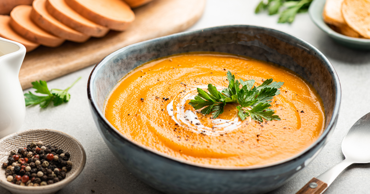 A bowl of sweet potato soup that's garnished with cream and parsley on a kitchen bench. Nearby is a dish of peppercorns, diced sweet potatoes, and a spoon.