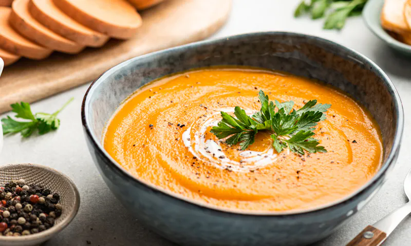 A bowl of sweet potato soup that's garnished with cream and parsley on a kitchen bench. Nearby is a dish of peppercorns, diced sweet potatoes, and a spoon.