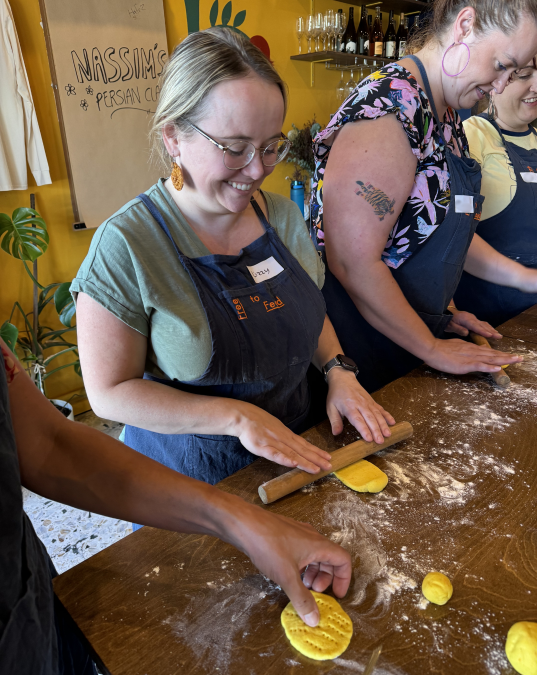 Lizzy is at a workbench in a cooking class, rolling out dough. She's is standing beside two other women, and they're all wearing aprons.