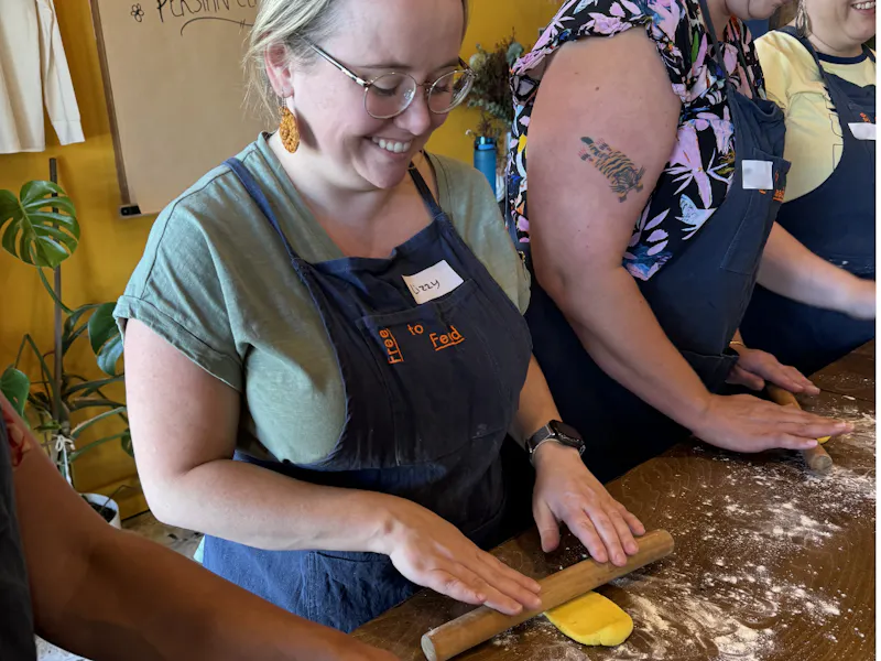 Lizzy is at a workbench in a cooking class, rolling out dough. She's is standing beside two other women, and they're all wearing aprons.