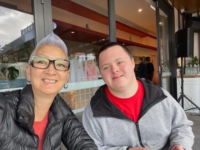 Jedd and his mum, Tina, are sitting outside at a cafe. Jedd has short dark hair and is wearing a grey jacket that's zipped up. Tina has short grey hair,  glasses, and has a puffer jacket on. They're both wearing jackets and are smiling gently. 