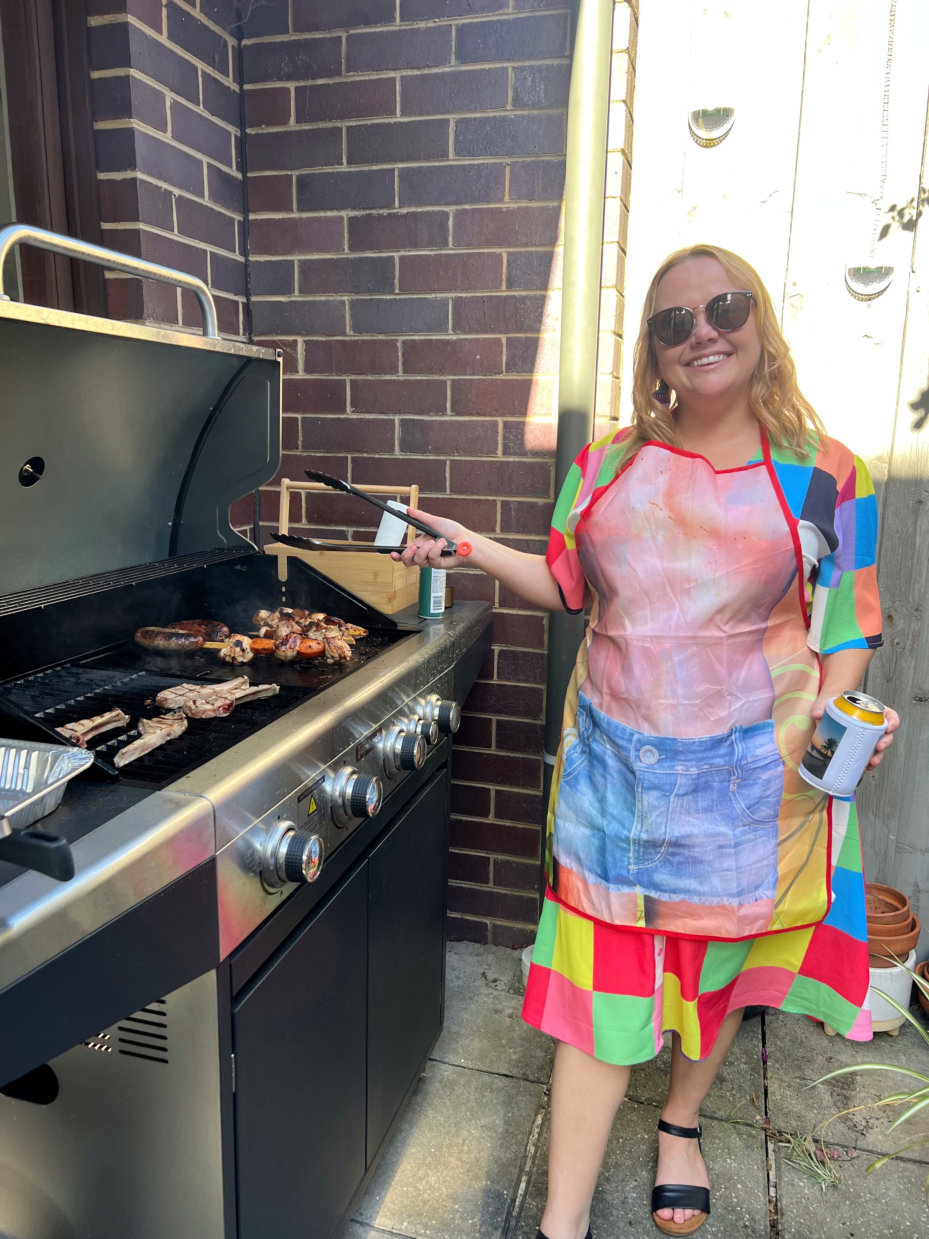Lizzy is standing near a barbecue and is holding onto tongs. She's wearing a bright coloured dress, sunglasses, and apron. 
