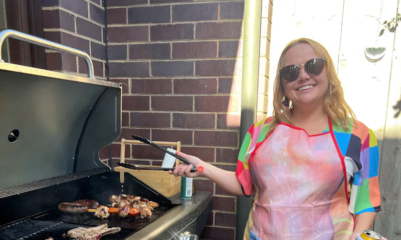 Lizzy is standing near a barbecue and is holding onto tongs. She's wearing a bright coloured dress, sunglasses, and apron.