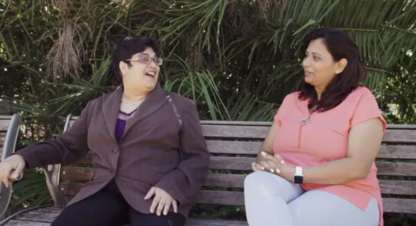 Nidhi and her Hireup Relationship Manager sit on a wooden bench outside. They are facing each other and smiling. Nidhi is wearing a brown jacket and her relationship manager is wearing a pink top.