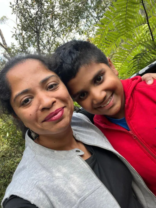 A smiling selfie photo outside of Shinoo and her son Aryansh. Shinoo wears a grey vest top and her black hair is tied back. Aryansh has black short hair and wears a red jumper.