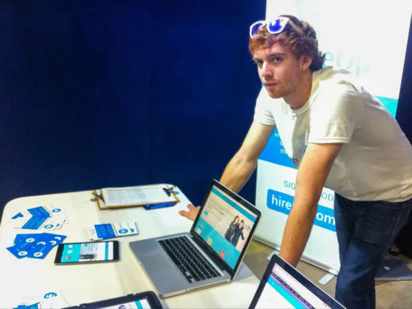 Hireup staff member, Ben Armstrong, is leaning on a table. He’s wearing a white t-shirt and has sunglasses perched on top of his head. Hireup information pamphlets are laid out on the table, while two laptops are open to Hireup’s first website.
