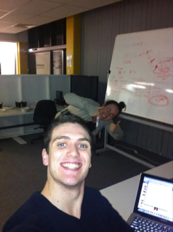 Jordan takes a smiling selfie in the Fighting Chance office. Laura O’Reilly is smiling in the background behind him giving a thumbs up. There is a large whiteboard with lots of ideas and writing in the background.