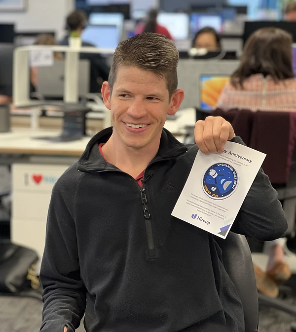 A smiling headshot of Westie in the Hireup office. He is holding up a Hireup patch and wears a black top.