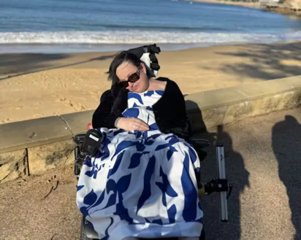A photo of Judy outside at the beach. She uses a wheelchair, dark sunglasses and wears a long blue and white dress.