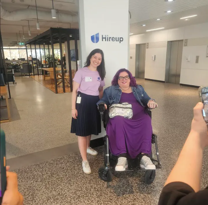 Two women are in an office building with the Hireup Logo on the wall in the background. On the left Maddy stands smiling at the camera. She is wearing a lavender colour tshirt, dark blue skirt and white sneakers. On the right V is using a wheelchair and is smiling at the camera. She is wearing a purple dress and a denim jacket with white sneakers.