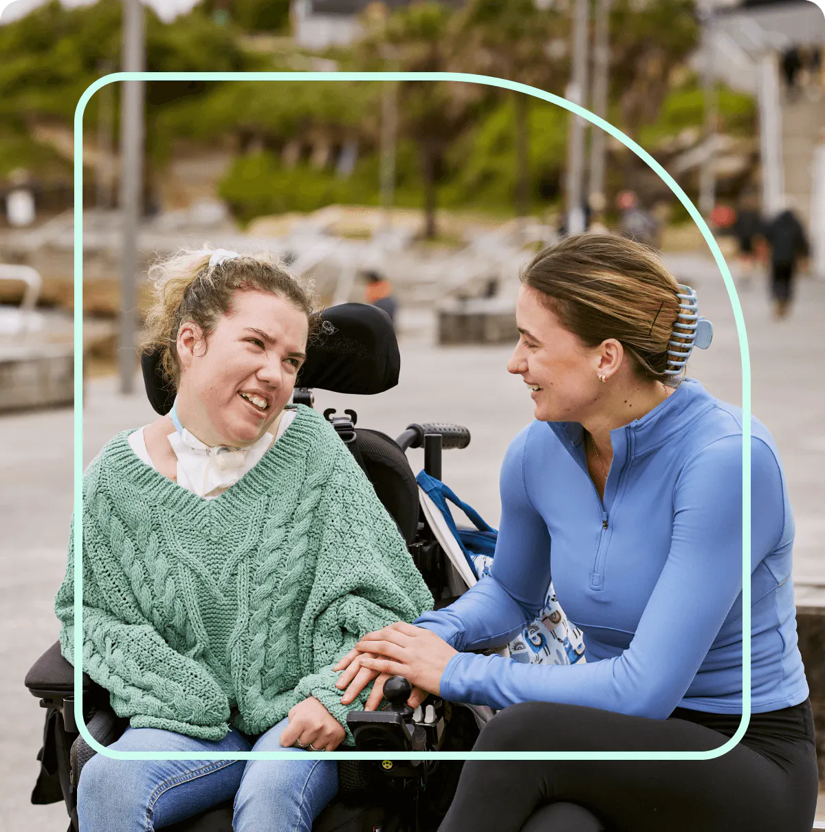 Two women are outdoors in a public space smiling at each other. The woman on the left uses a wheelchair and is wearing a green jumper and jeans. The woman on the right is sitting next to her wearing a blue top and black pants.