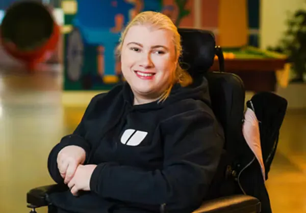 A smiling photo of Samantha Vella in the Hireup office. She is using a black wheelchair and wears a black hoodie with the Hireup logo. Her hair is blonde and tied back.