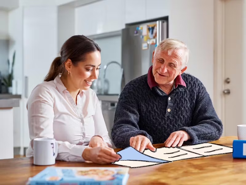 Dirk, a Hireup Aged Care client, is sitting at a dinning room table with his support worker, Brenda. Dirk has short, grey hair and is wearing a knitted jumper, while Brenda is wearing a white blouse. They're drinking coffee and are flipping through an illustrated newspaper.