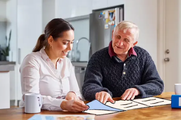 Dirk, a Hireup Aged Care client, is sitting at a dinning room table with his support worker, Brenda. Dirk has short, grey hair and is wearing a knitted jumper, while Brenda is wearing a white blouse. They're drinking coffee and are flipping through an illustrated newspaper.