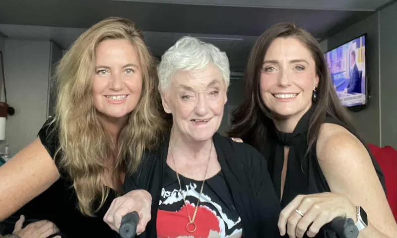 Narelle and her two daughters stand side by side, smiling. They're in a hotel room and are dressed in evening clothes. Narelle is holding onto a walker.