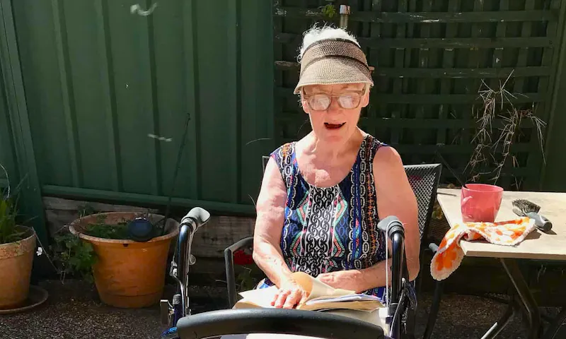 Narelle, a Hireup aged care client, is sitting outside in the sunshine. She's wearing a visa and short-sleeved top. Narelle's cat, Will, is standing near her walker.