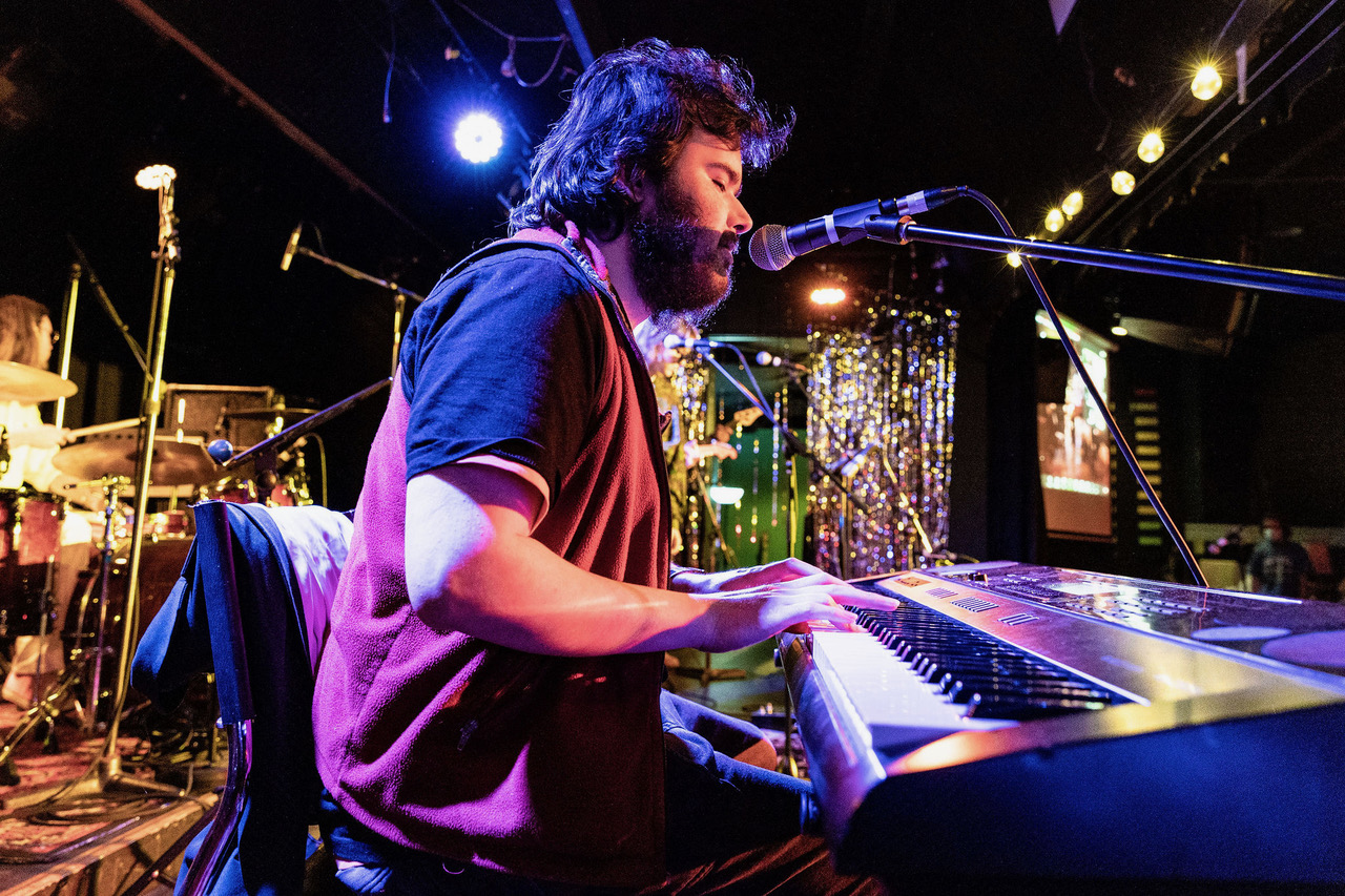 Edward, a neurodivergent music graduate, is playing a piano keyboard on a stage. A spotlight is shining on him and he has dark, woolly hair and beard. 