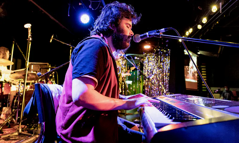 Edward, a neurodivergent music graduate, is playing a piano keyboard on a stage. A spotlight is shining on him and he has dark, woolly hair and beard.