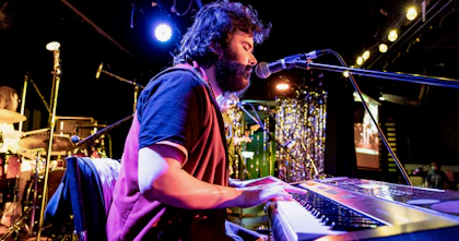 Edward, a neurodivergent music graduate, is playing a piano keyboard on a stage. A spotlight is shining on him and he has dark, woolly hair and beard.