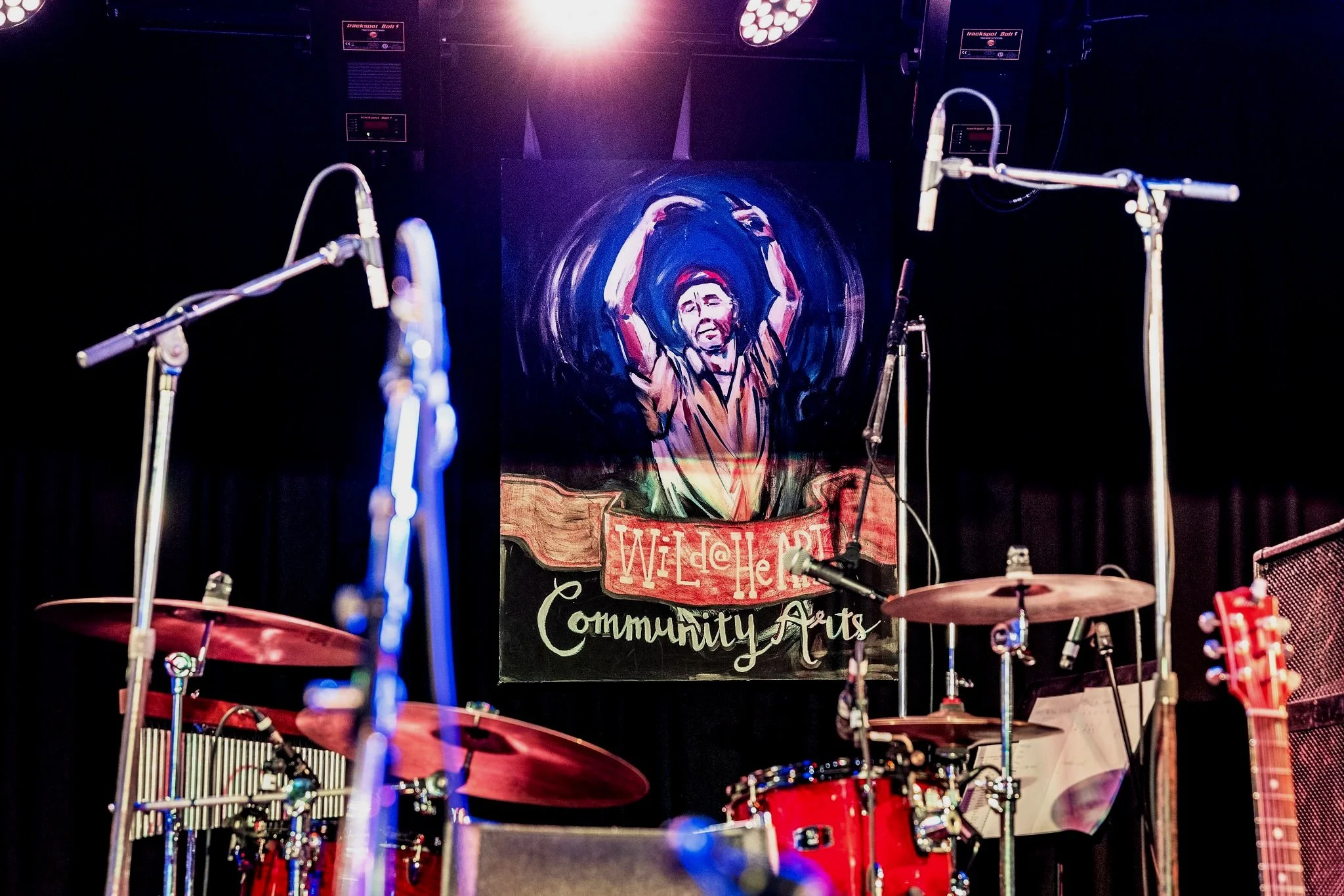 A poster of Wild at Heart, an inclusive music community group, features a man dancing. The poster is the backdrop to a stage that has guitars and a drum set.