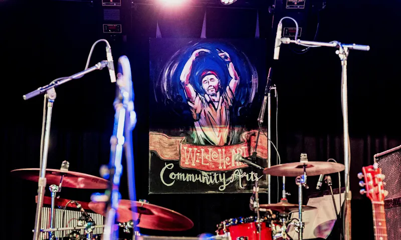 A poster of Wild at Heart, an inclusive music community group, features a man dancing. The poster is the backdrop to a stage that has guitars and a drum set.
