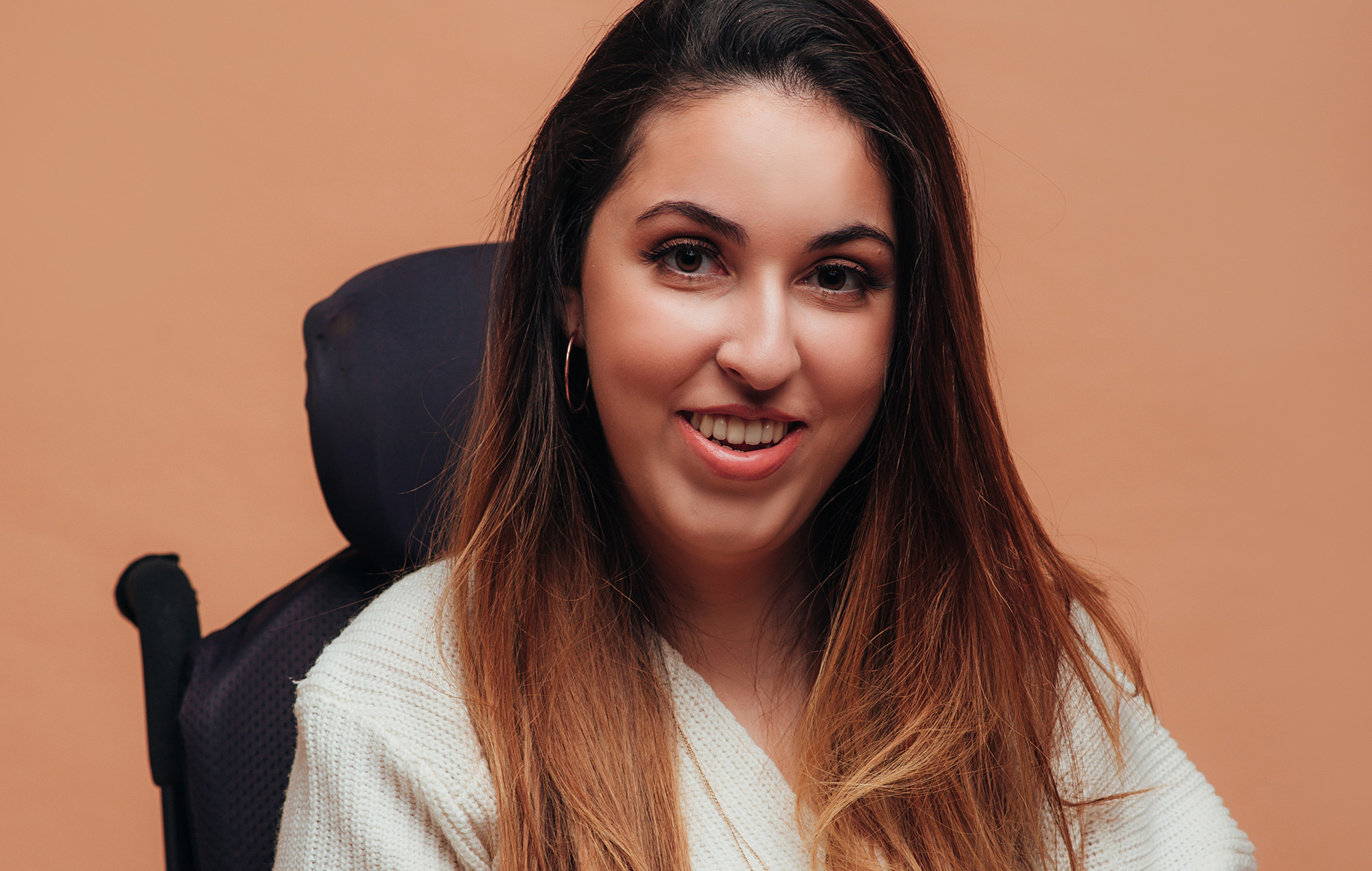Dina Bassile, founder of Tibi Access, is smiling gently, has long hair and is wearing a white jumper. Dina is a wheelchair user.