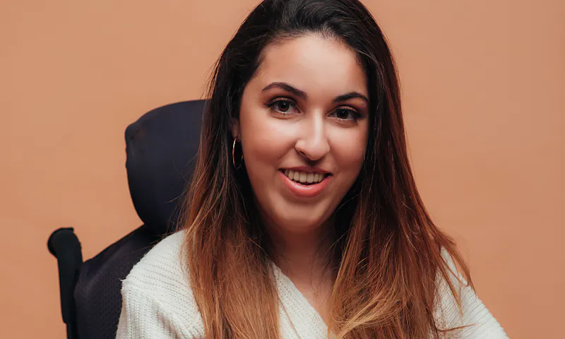 Dina Bassile, founder of Tibi Access, is smiling gently, has long hair and is wearing a white jumper. Dina is a wheelchair user.