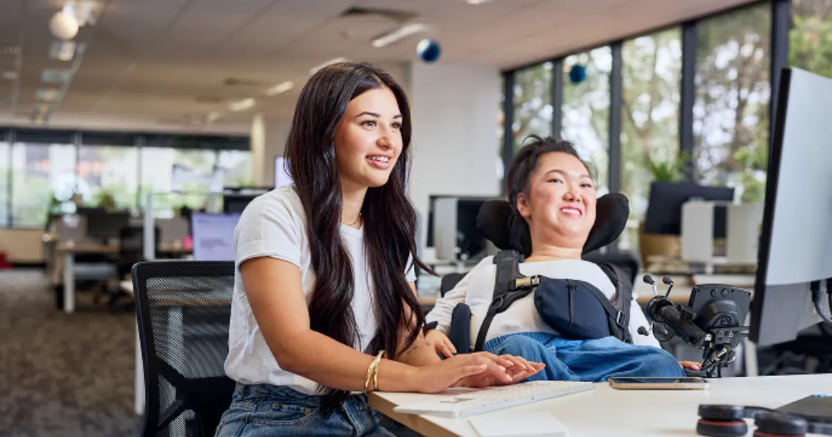 Melanie and her support worker Allyah are in an office. They're both wearing white t-shirts, jeans, and are smiling. Allyah is mid-typing on a desktop computer.