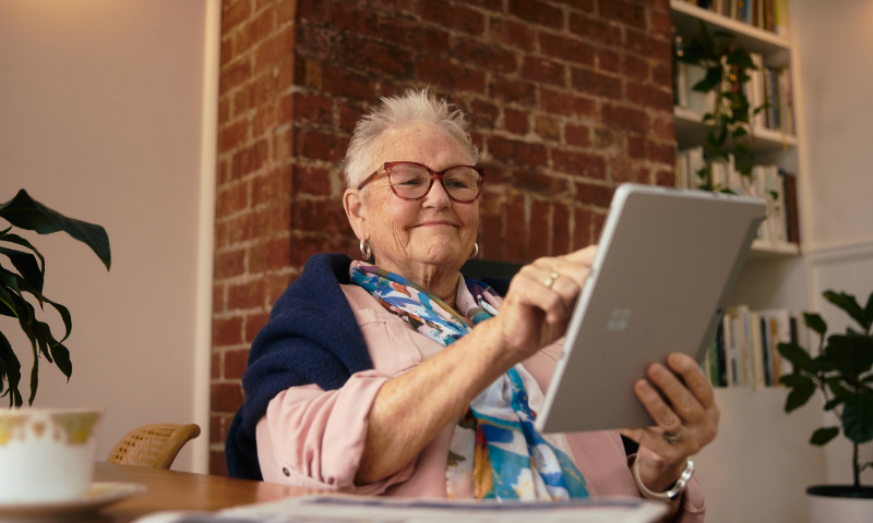 A woman is tapping an iPad. She's sitting at a dining table and wears glasses, a pink top, scarf, and has a cardigan draped over her shoulders.