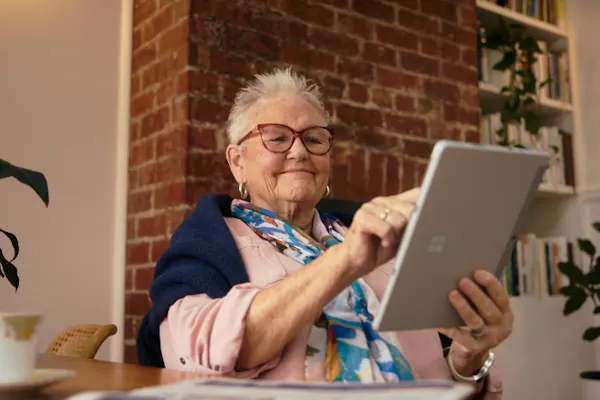 A woman is tapping an iPad. She's sitting at a dining table and wears glasses, a pink top, scarf, and has a cardigan draped over her shoulders.