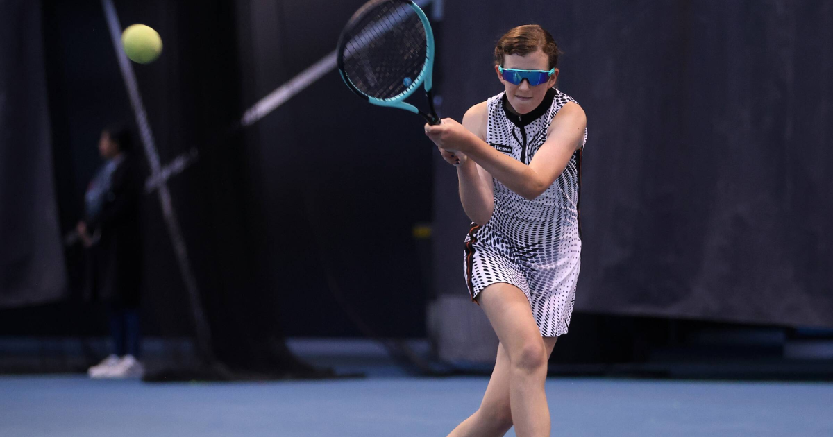 Maddy Kelly whacks a tennis ball across a blue court. Maddy, who is Australia's top-ranked junior female Blind and Low Vision tennis player, wears sunglasses and a tennis dress. 