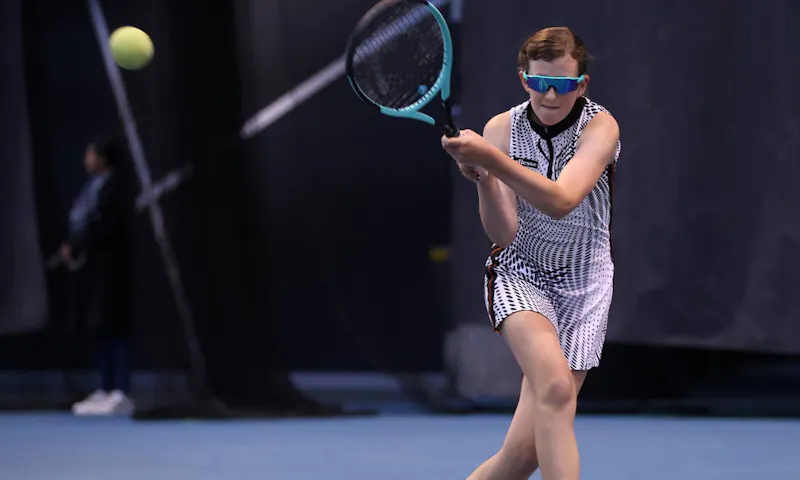 Maddy Kelly whacks a tennis ball across a blue court. Maddy, who is Australia's top-ranked junior female Blind and Low Vision tennis player, wears sunglasses and a tennis dress.