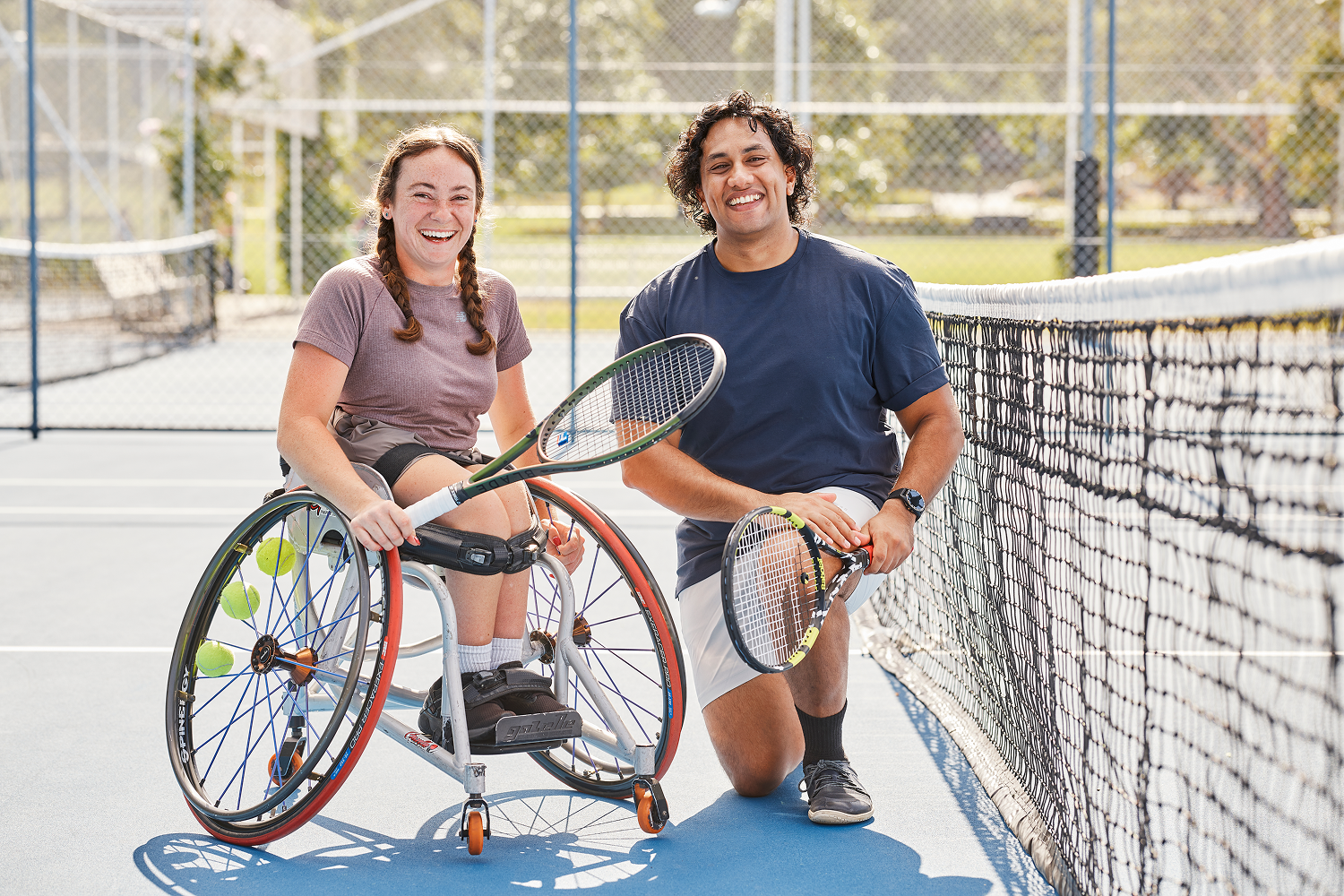 Hireup client Hayley holds a tennis racket and smiles with her support worker Shay on a tennis court.