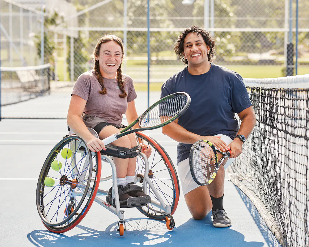Hireup client Hayley holds a tennis racket and smiles with her support worker Shay on a tennis court.