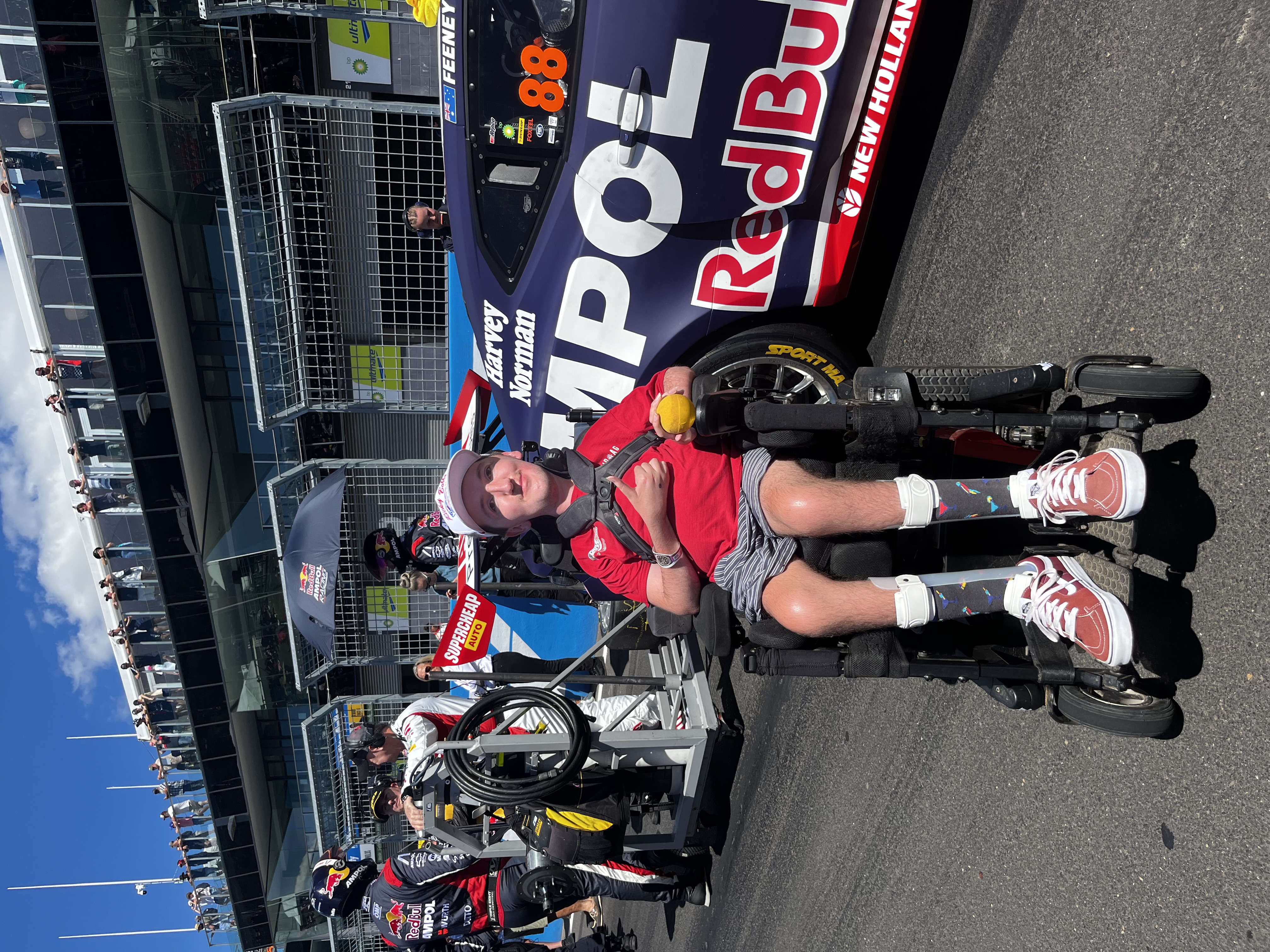 Tom Giumelli is giving a 'thumbs-up' beside a race car that's parked in pit lane. Tom has short hair and is wearing a cap, t-shirt and shorts. He also wears AFOs (Ankle Foot Orthotics) and is a wheelchair user.