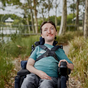 Tom is a twenty-something-year old man and is pictured on a grassy footpath near a lake. He has short, dark hair and wears a teal coloured top and jeans. Tom's a wheelchair user and is wearing a chest harness.