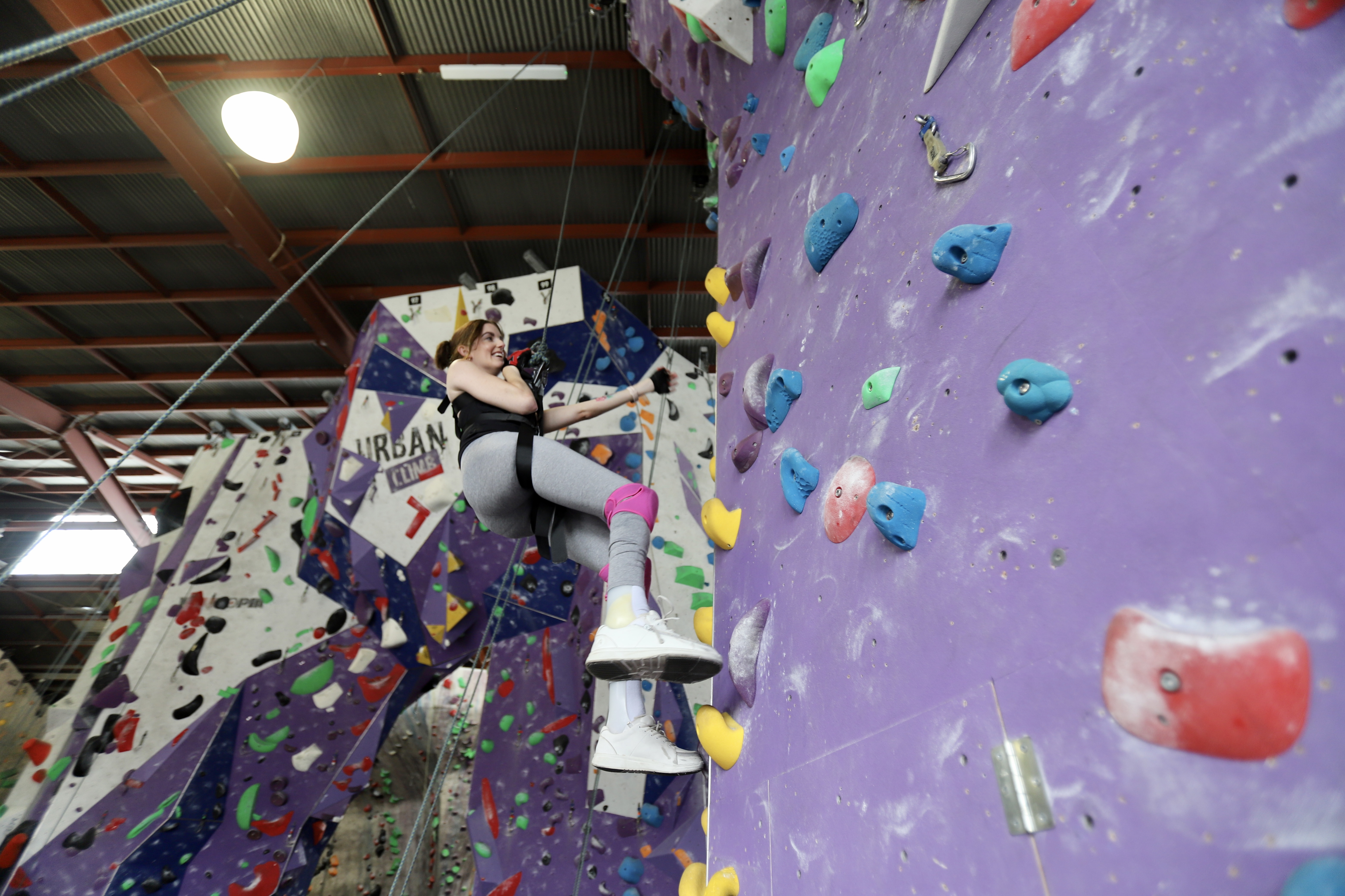 Sam swings on ropes at a rock climbing gym. She's reaching out to grasp the wall which is covered in rocky bumps. Sam is smiling. 