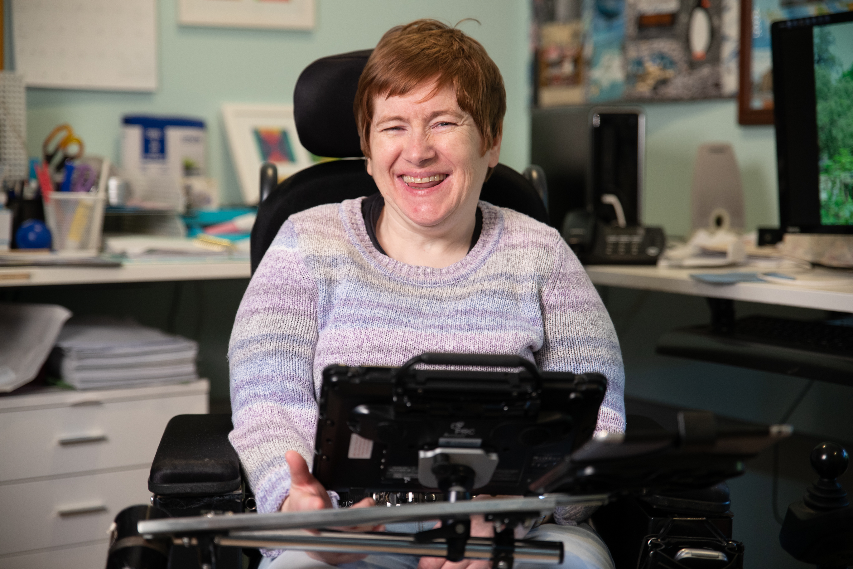 Fiona has red hair and is wearing a purple jumper. Behind her is a home office setting with a long desk, computer desktop and stationary. Fiona is a wheelchair user and has an Alternative and augmentative communication device in front of her. Fiona is smiling. 