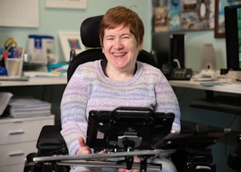 Fiona has red hair and is wearing a purple jumper. Behind her is a home office setting with a long desk, computer desktop and stationary. Fiona is a wheelchair user and has an Alternative and augmentative communication device in front of her. Fiona is smiling.