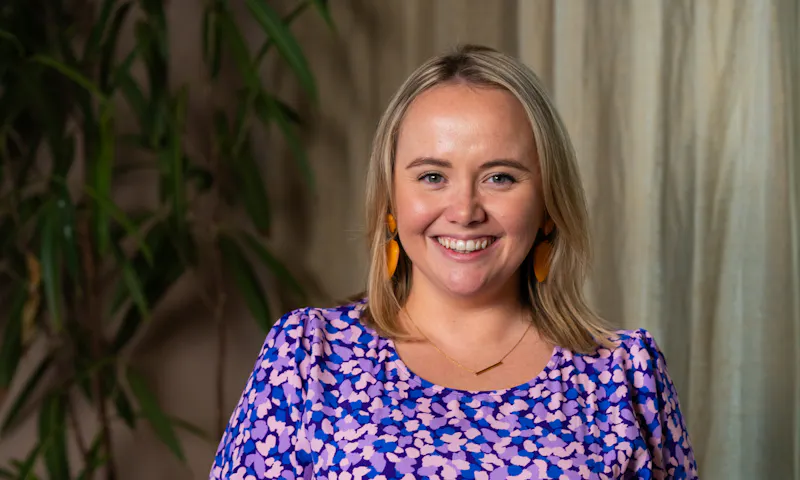 Lizzy is smiling. She has short blonde hair and is dressed in a purple polka dot dress, complemented by orange earrings.