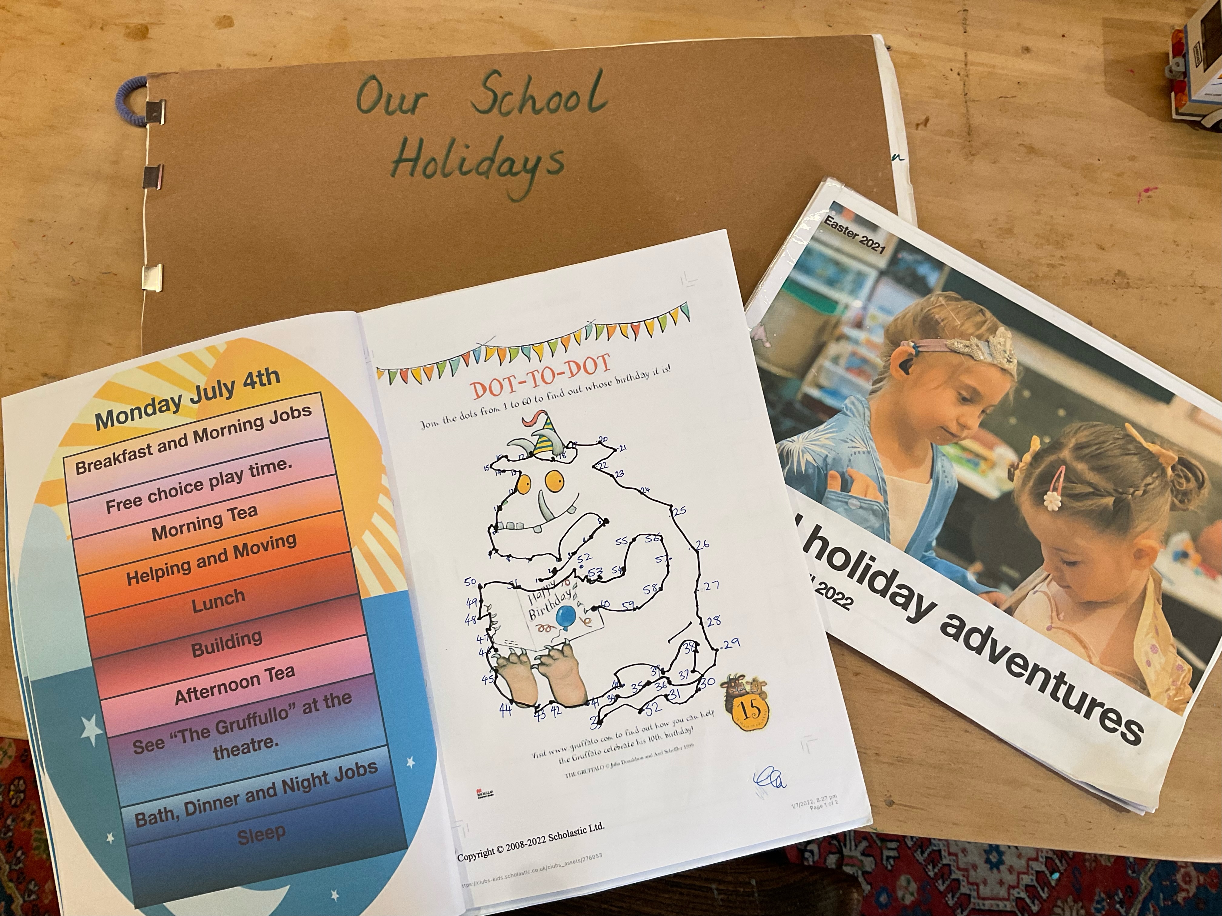 Three colourful activity booklets are laid out on a table. The titles include 'Our School Holidays', 'Holiday adventures' and 'dot-to-dot' task. 