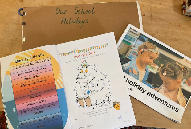 Three colourful activity booklets are laid out on a table. The titles include 'Our School Holidays', 'Holiday adventures' and 'dot-to-dot' task.
