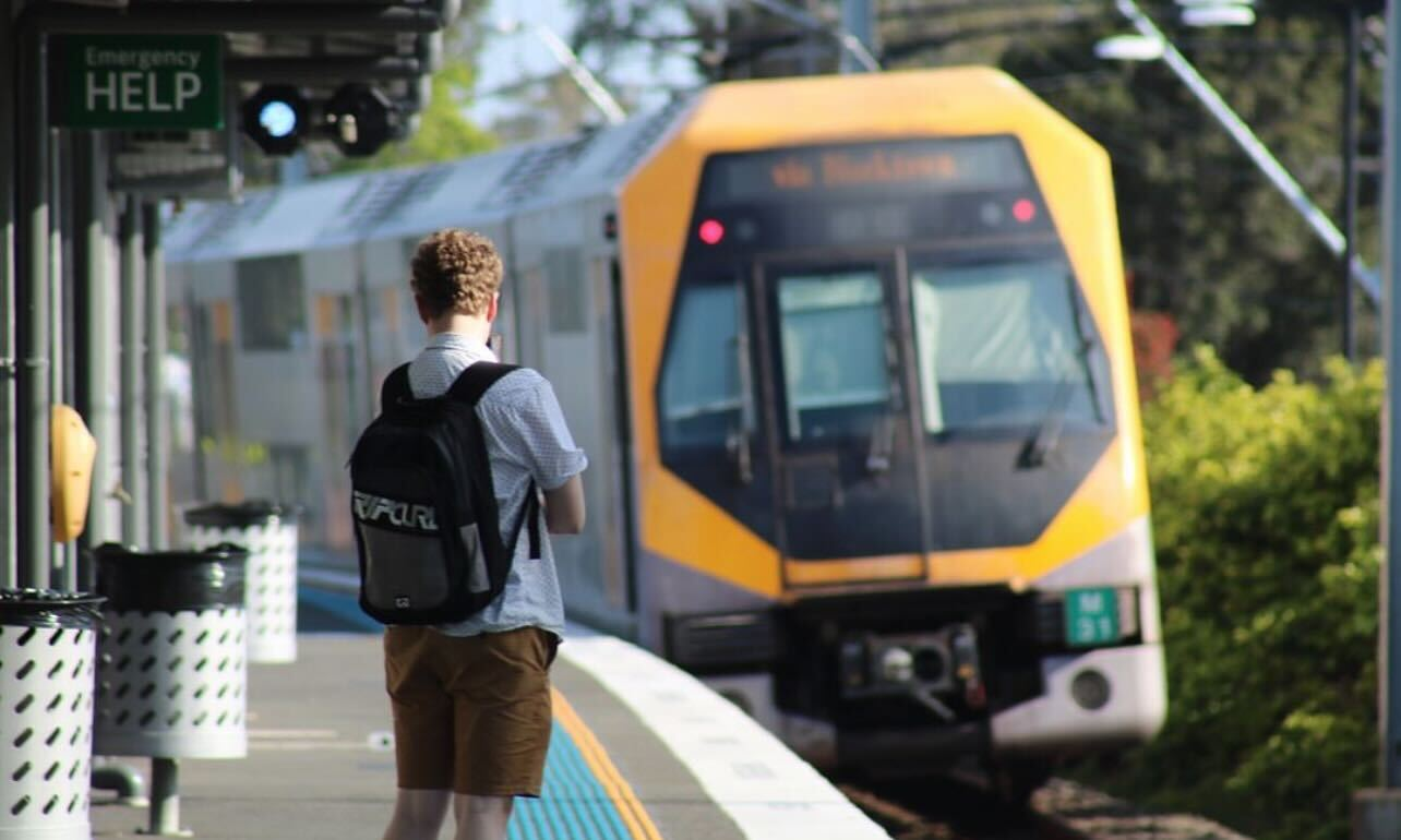 Julian is standing on a train platform in Sydney. He has short curly blonde hair and is wearing a black Ripcurl backpack He is wearing a white collared shirt and brown shorts. In the background a yellow and silver train is approaching the platform.