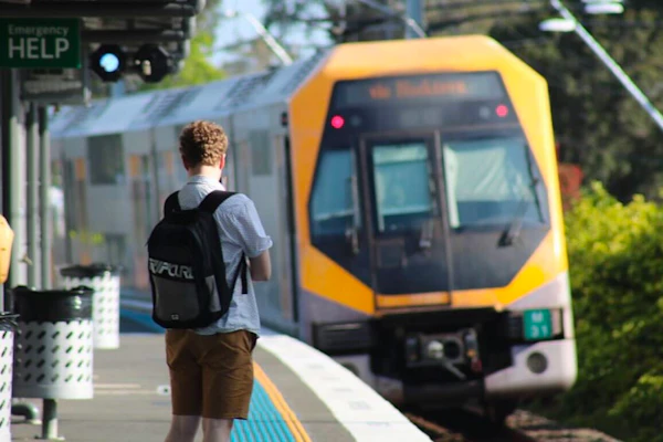 Julian is standing on a train platform in Sydney. He has short curly blonde hair and is wearing a black Ripcurl backpack He is wearing a white collared shirt and brown shorts. In the background a yellow and silver train is approaching the platform.